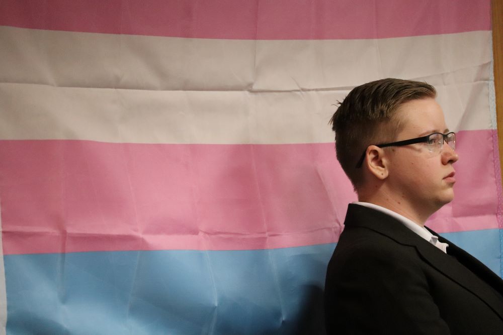 A volunteer stands alone along side the trans flag. Photo credit: Mary Vogel