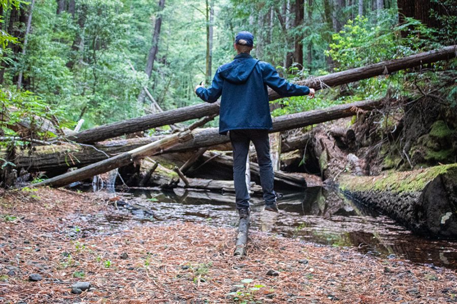 A man balancing on one leg on a narrow log above a creek.