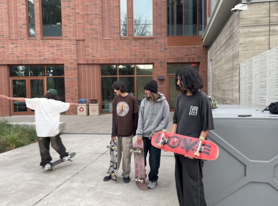 three boys holding skateboards in front of a brick building while another boy does a skateboard balance trick next to them