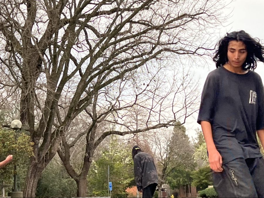 Teenager outside wearing all black. Leafless trees behind him as well as another teen farther back