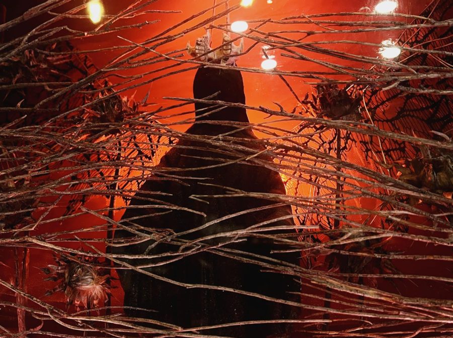 Multiple thin sticks stretch out in front of a heavily shadowed idol. Small dead flowers frame the idol as dark red lights are static in the background.