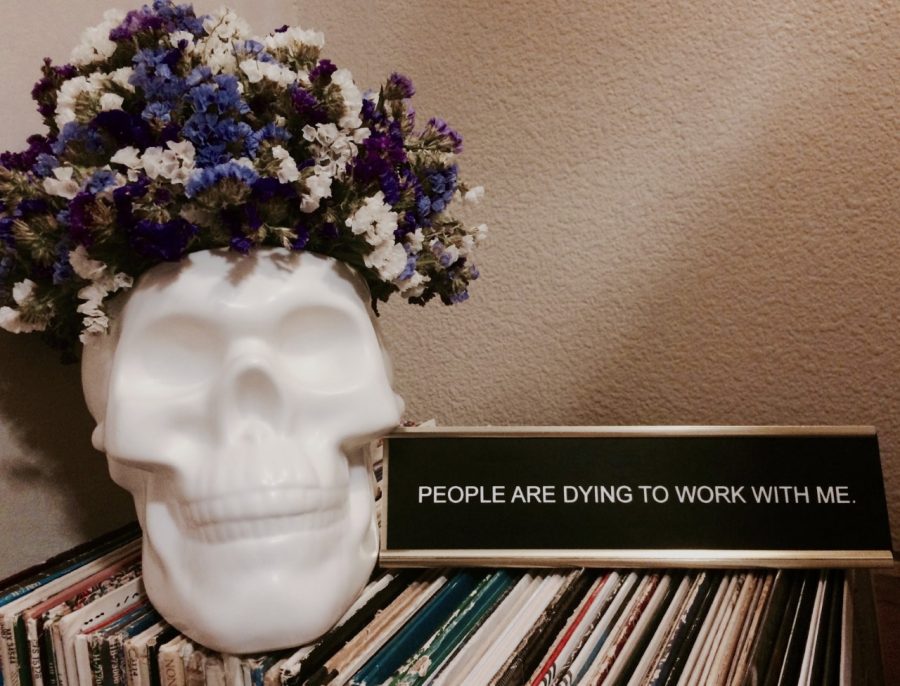 A skull vase full of blue, white and purple flowers sits next to a sign that reads "people are dying to work with me."