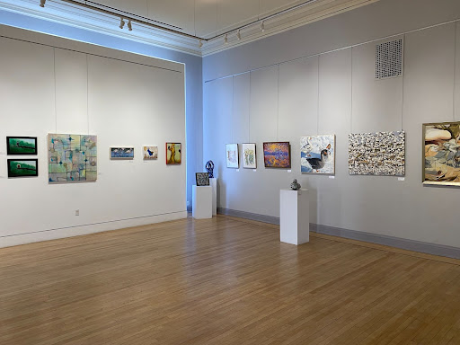 open spacious room with white walls and light brown hardwood floors. Colorful abstract and nonobjective art hangs on the walls in a line. Three rectangular pedestals display art.