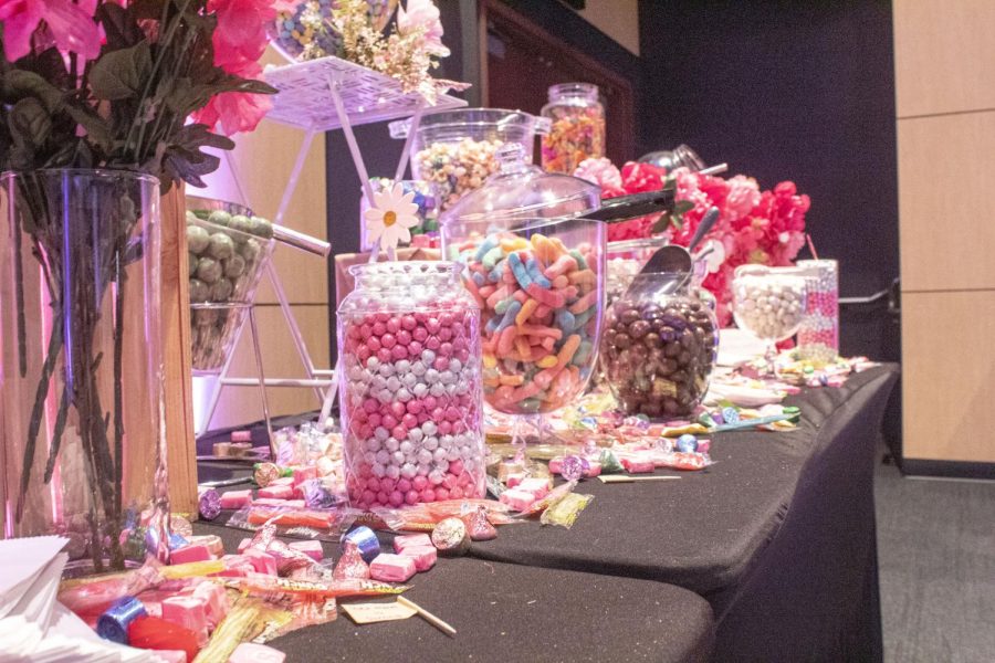 Eight jars of different types of candy sit on a black clothed table, surrounded by cases of pink and red flowers.