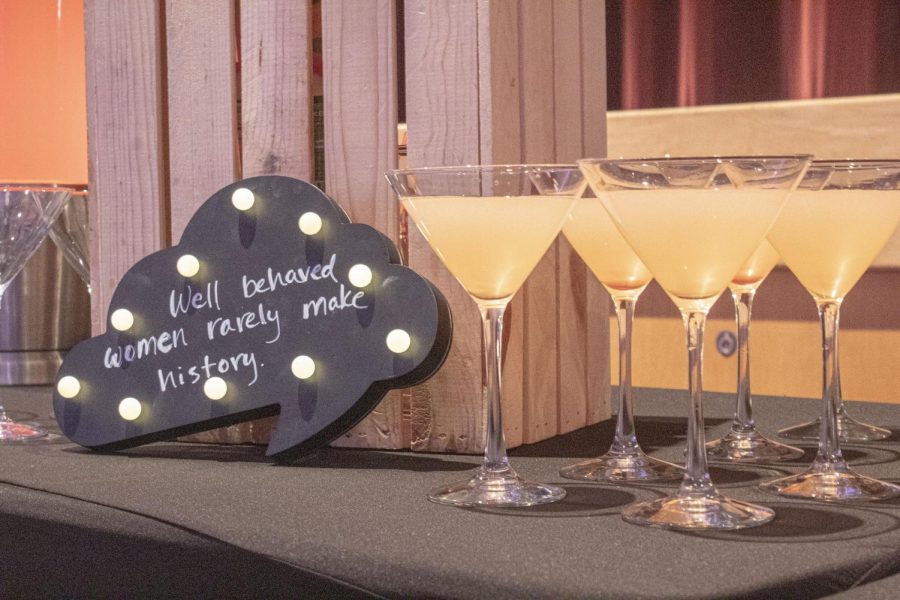 A cloud-shaped black sign that says "Well behaved women rarely make history" sits next to five pink-tinted mocktails in martini glasses.