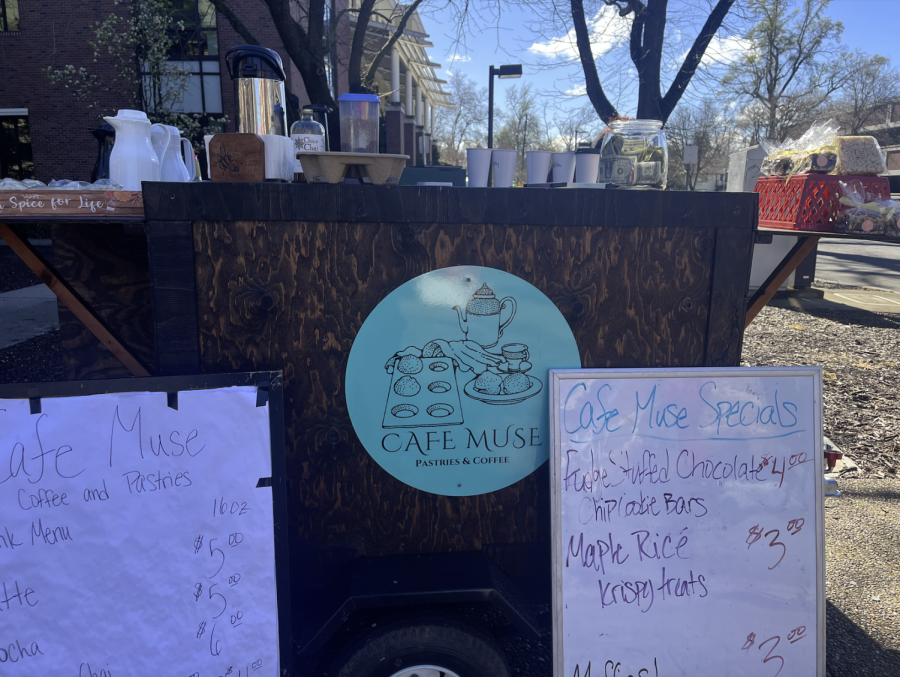 A dark wood coffee cart sits at an intersection. A circular, cyan emblem sits attached to the middle of the front of the cart. Two whiteboards sit on either side of the emblem.
