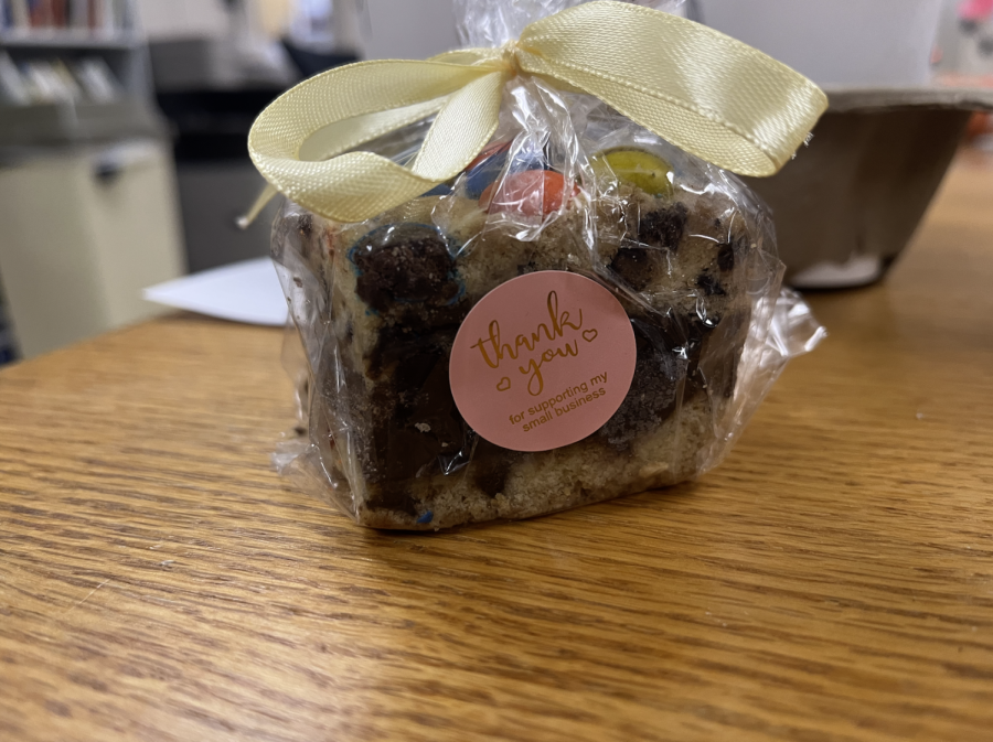 Cafe Muse's fudge stuffed chocolate chip cookie bar sits inside of translucent packaging tied off by a golden ribbon. A light pink circular sticker says "Thank you for supporting my small business" in gold metallic lettering.
