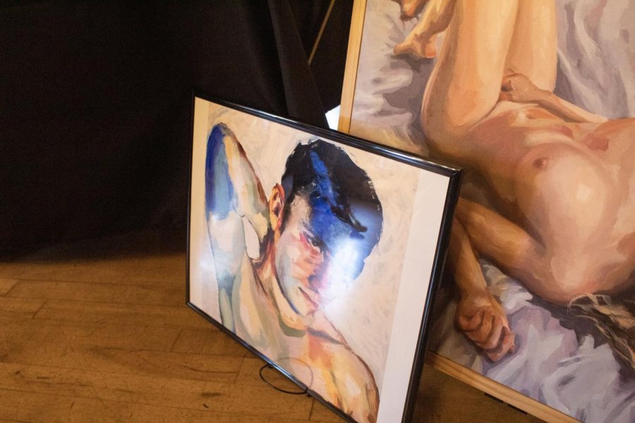A blue and naturally colored painting sits in front of a larger painting of a naked person laying on a sheet.