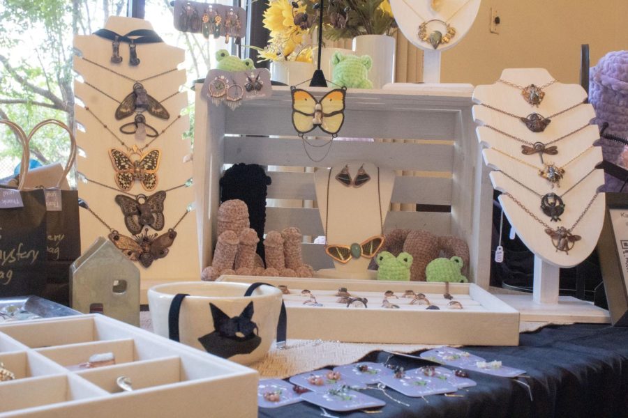 A multitude of nature themed penis and nude bust pendants, necklaces and earrings sit on display next to a white crate box filled with crocheted penises, vaginas and frogs.