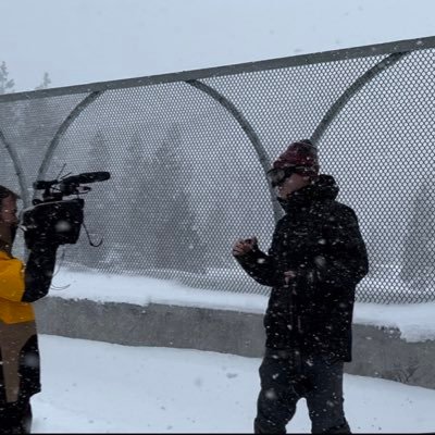 man in all black with goggles and hat in snow on a bridge being filmed by a man with a large camera who is wearing a yellow jacket