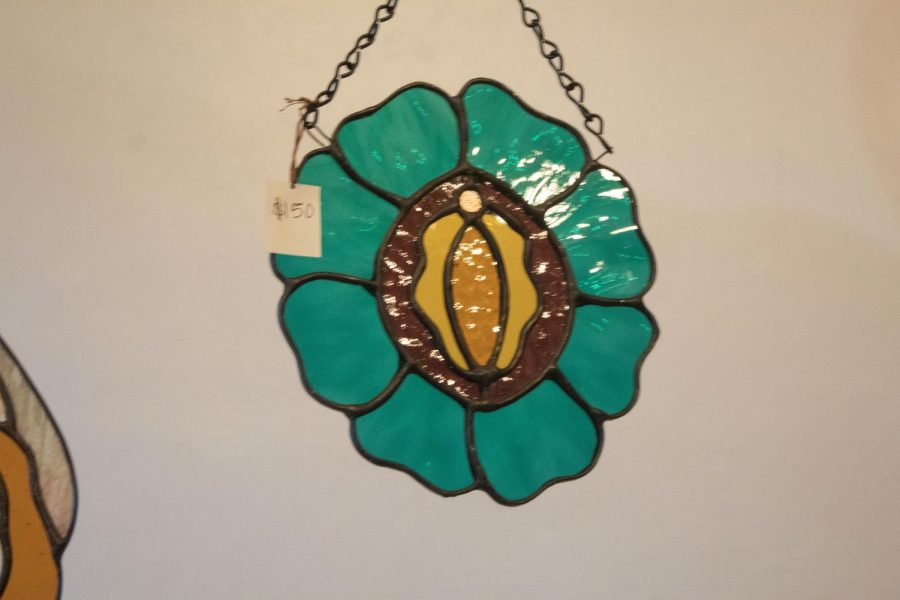 A vulva is centered inside of a teal flower shaped stained glass hanging.
