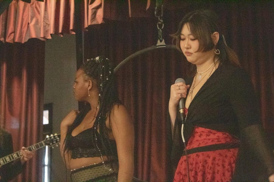 Vocalist Demondra Martin looks to the left toward Susana Correa Avila Robb as she sings. Alexis Ong looks down at the stage as she holds the wired microphone class to her chest in one hand.