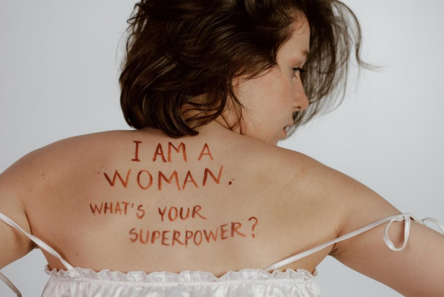 A woman looks over her shoulder. I am a woman. Whats your superpower? is written in dark red paint on the models back. Shes wearing a white tank top, the straps sliding off of her shoulders.