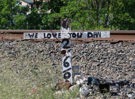 White cross with black hand-painted text reading We love you Dan is placed in gravel in front of railroad tracks.
